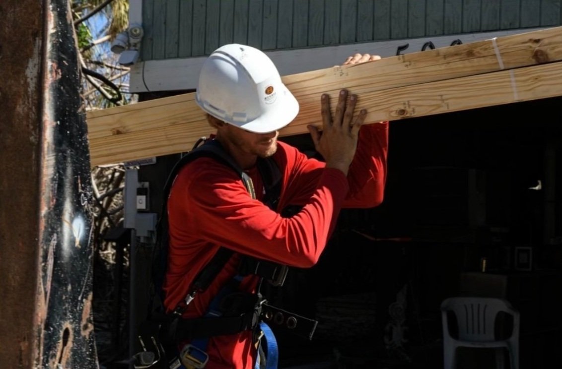 The same job, lumber on the shoulder, a damaged house behind him.