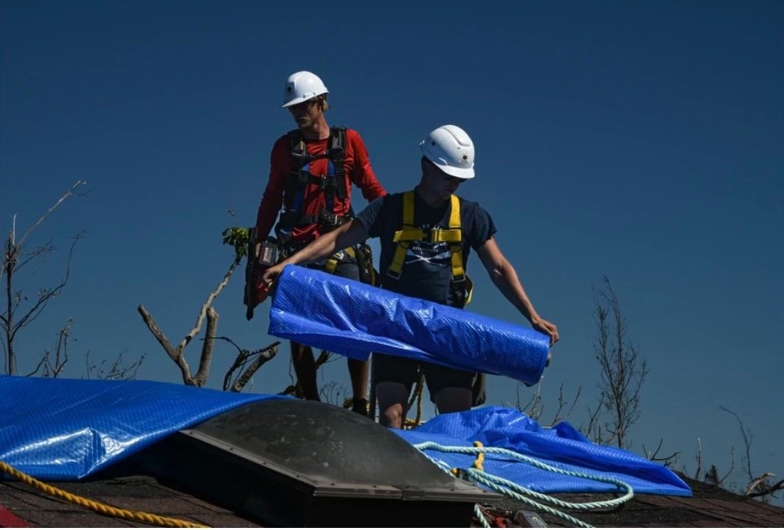 Two crew members on a roof unrolling a blue tarp after a storm.
