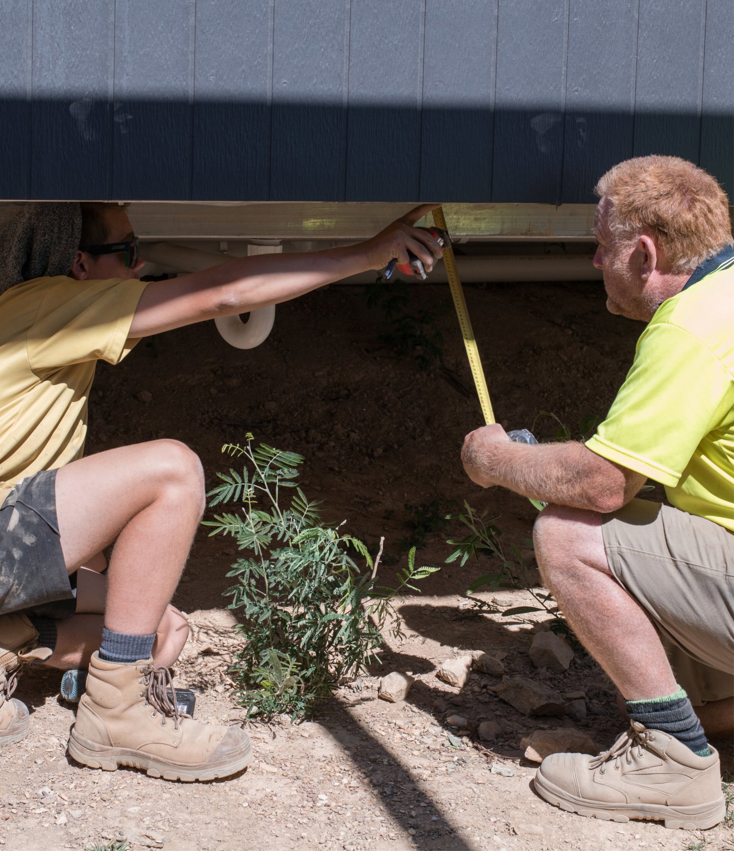 Two people measuring under a house on a crawlspace job.